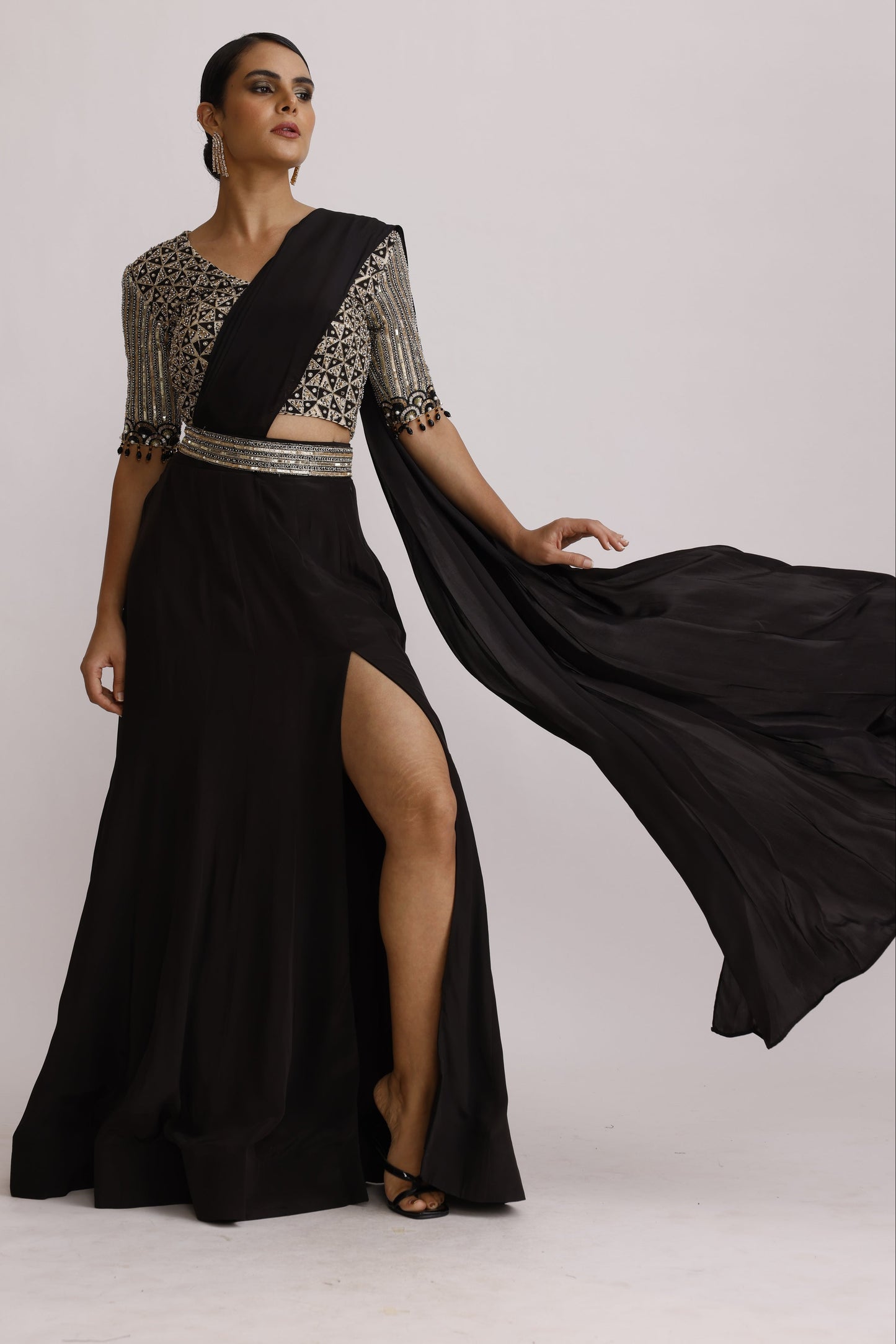 A woman modeling a black floor-length gown with a sheer draped overlay and heavily embellished silver beaded sleeves, posed against a plain light background.