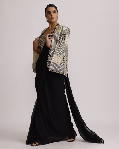 A woman models a black floor-length dress with a long flowing train, styled with a beige and black patterned embroidered jacket. She is standing against a solid light gray studio background.