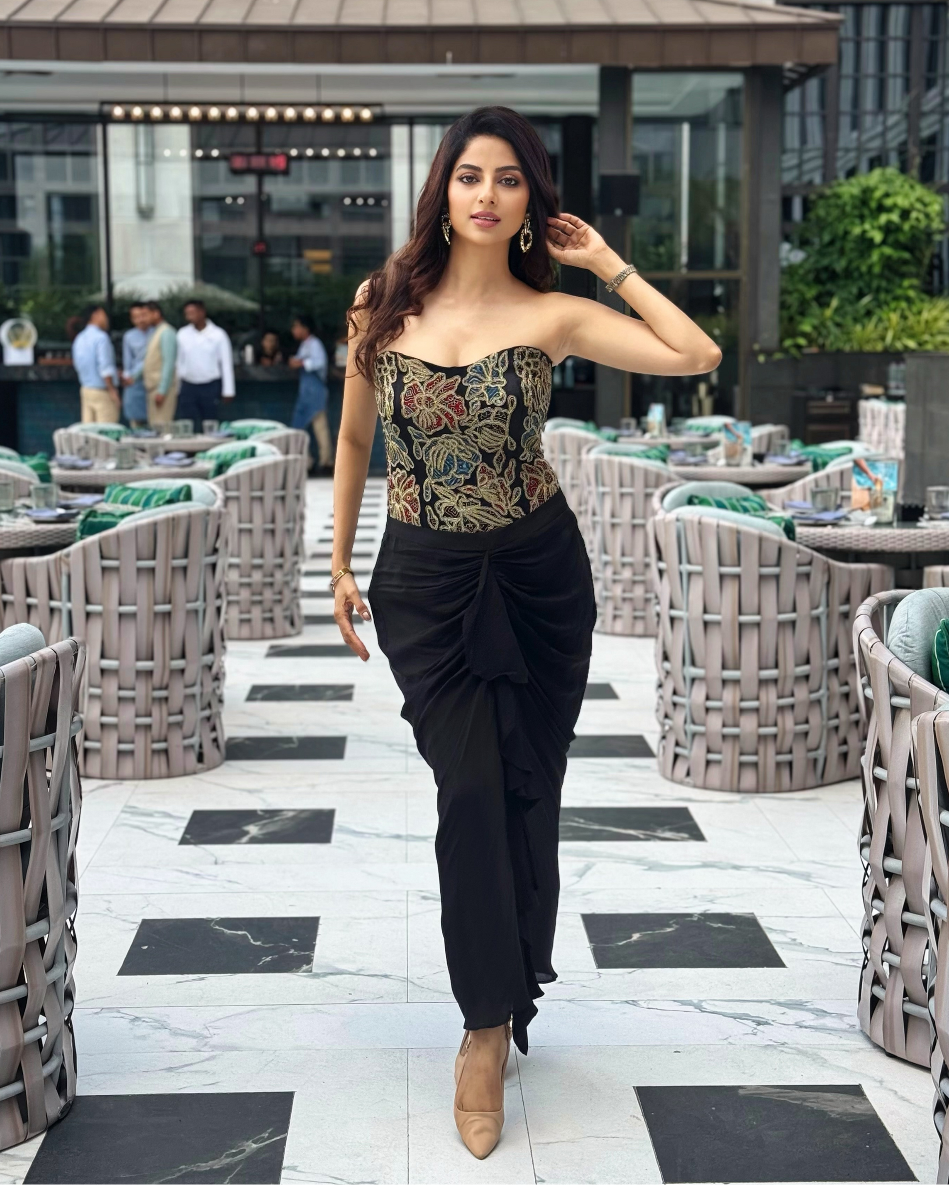 A Ekta Maru posing on an outdoor patio walkway, wearing a strapless corset-style top with a multicolor paisley pattern and black draped trousers.