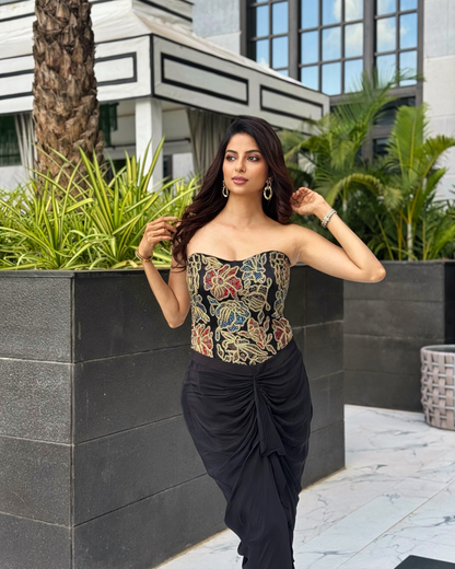 A Ekta Maru models a strapless corset-style top with an intricate multicolor floral embroidery pattern on a black base, paired with a black draped wrap skirt, set against a modern outdoor patio background with green plants.