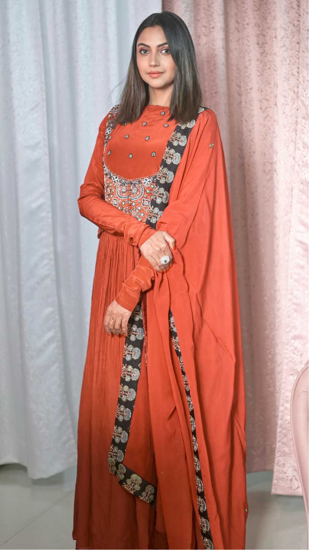 A Falak Khan wearing a traditional Indian outfit consisting of an orange-red anarkali kurta with silver embroidery on the chest, matching wide-leg pants, and a draped dupatta with a black border featuring gold and silver floral patterns. She is standing in front of a white and pink curtain backdrop.