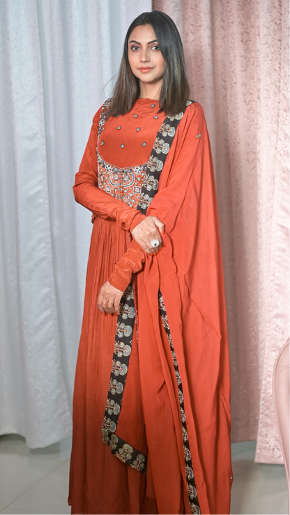 A Falak Khan wearing a traditional Indian outfit consisting of an orange-red anarkali kurta with silver embroidery on the chest, matching wide-leg pants, and a draped dupatta with a black border featuring gold and silver floral patterns. She is standing in front of a white and pink curtain backdrop.