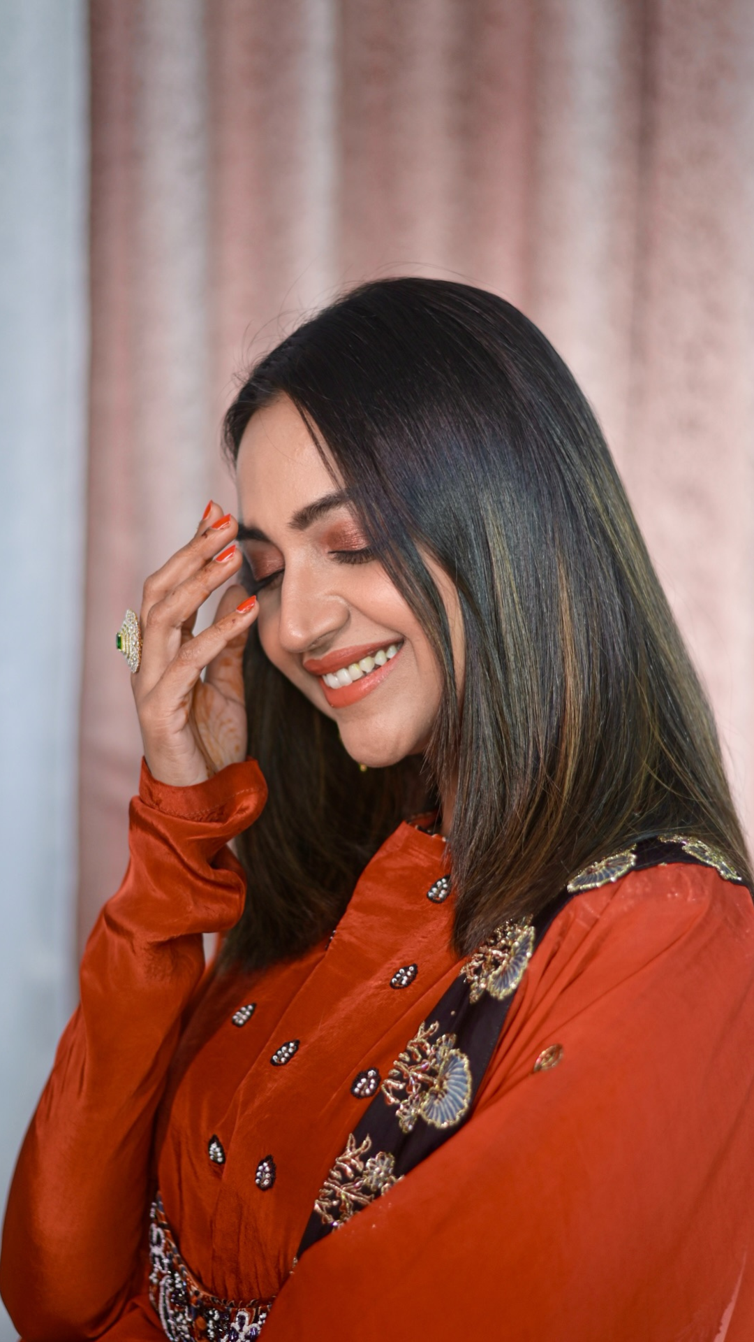 A Falak Khan with long dark hair is shown in a close-up portrait, wearing a rust-orange embroidered traditional outfit against a soft pink curtain background.