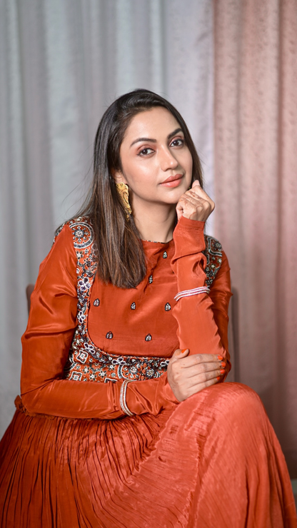 A Falak Khan wearing a rust-orange traditional outfit with embroidered detailing on the bodice, seated against a soft draped curtain backdrop.