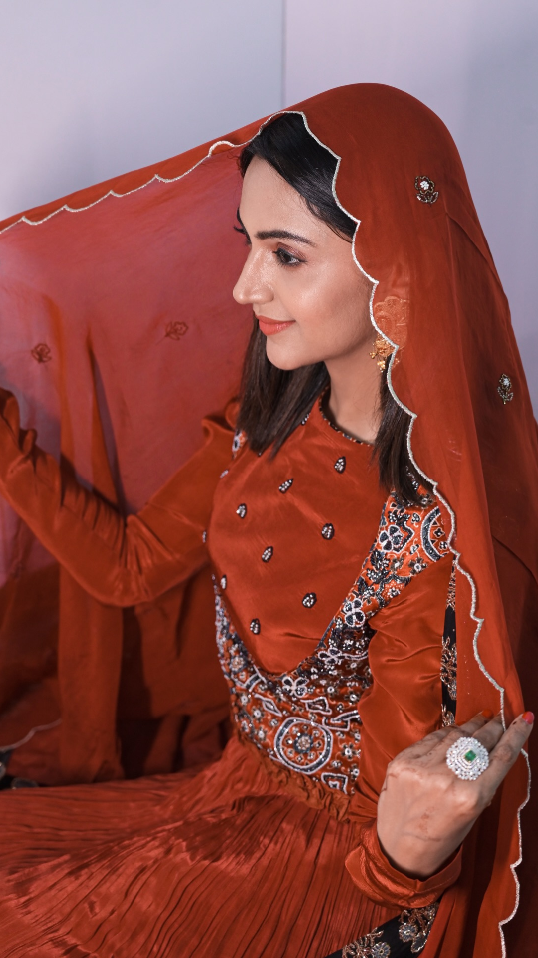 A Falakh Khan wearing a traditional rust-orange embroidered dress with a matching sheer dupatta draped over her head, posed against a light gray background.