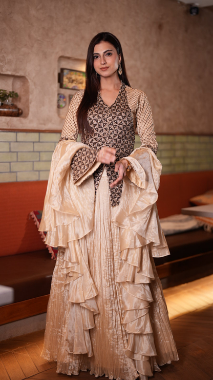 A Farhana Bhatt modeling a traditional Indian outfit with a beige and gold tiered skirt, a patterned brown and gold blouse, and dramatic bell sleeves in a warm indoor setting.