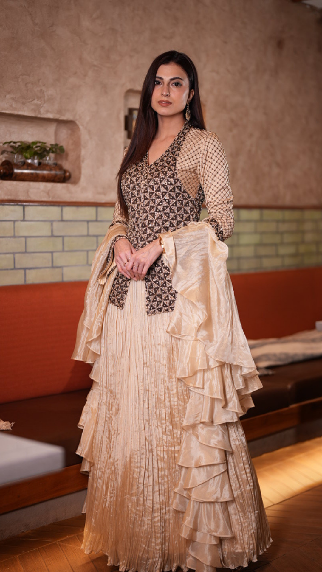 A Farhana Bhatt modeling a traditional Indian outfit featuring a beige and brown patterned kurta with a matching tiered dupatta, set against an indoor background with a textured wall and wooden furniture.
