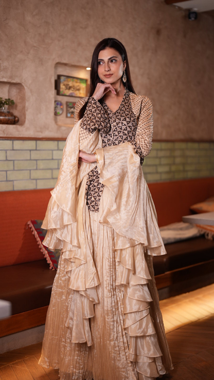 A Farhana Bhatt models a traditional Indian outfit consisting of a beige and brown embroidered kurta with a matching tiered pleated skirt, set against an indoor background with a textured wall and wooden bench.