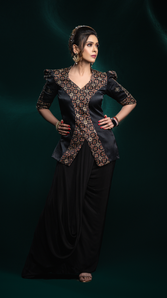 A Hrishitaa Bhatt models a black traditional outfit with ornate gold and brown embroidery on the bodice and sleeves, paired with a long black skirt, set against a dark green studio background.