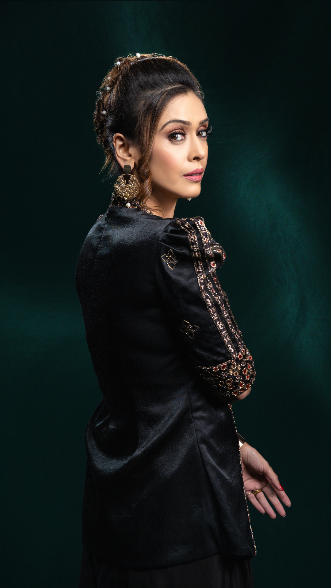 A Hrishitaa Bhatt in a black traditional outfit with ornate gold embroidery on the sleeve, photographed against a dark green studio background.