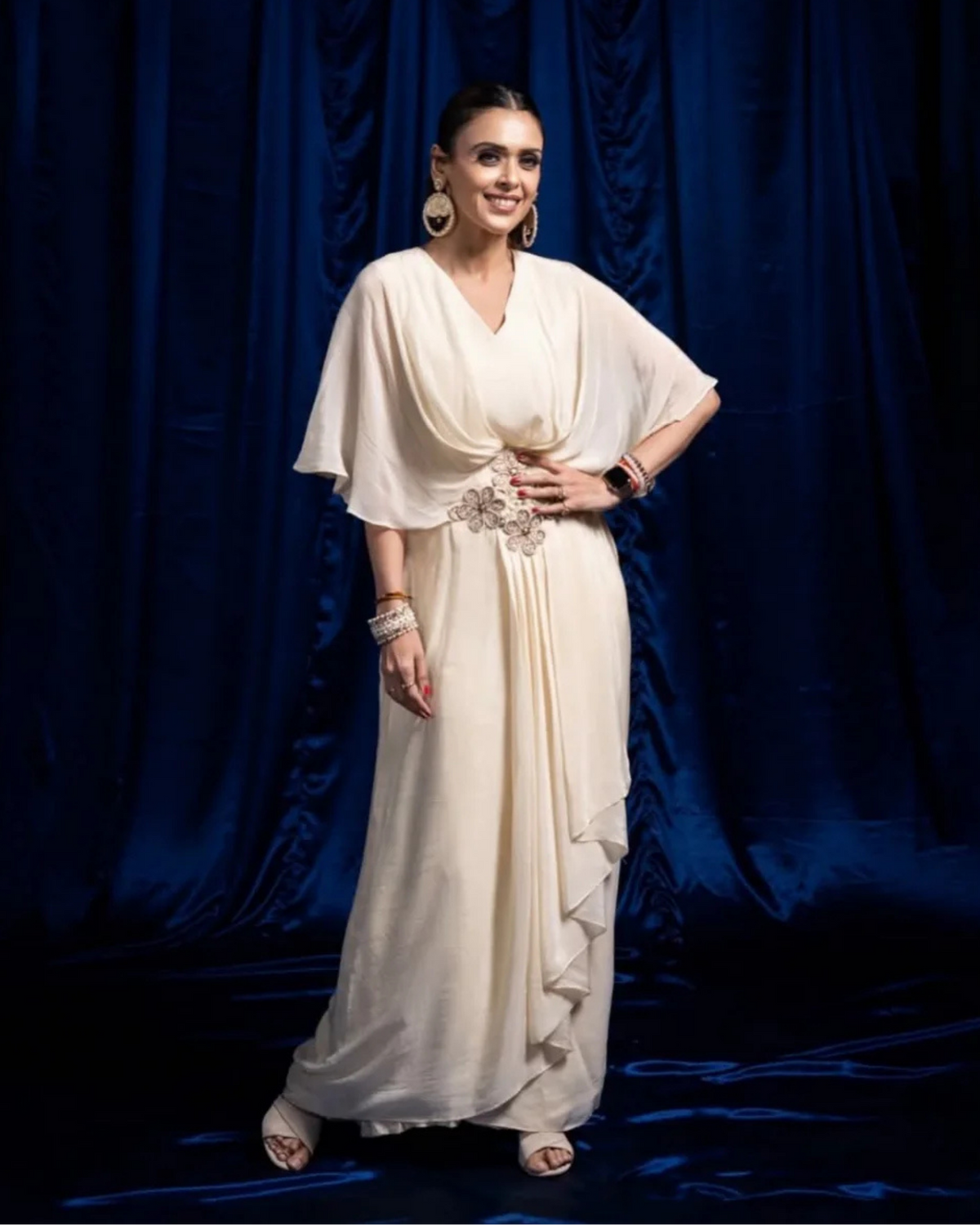 A Hrishitaa Bhatt stands in front of a dark blue draped curtain wearing a flowing ivory formal gown with draped sleeves, a gathered waist, and layered ruffle details along the skirt. She is also wearing statement earrings, bracelets, and sandals.