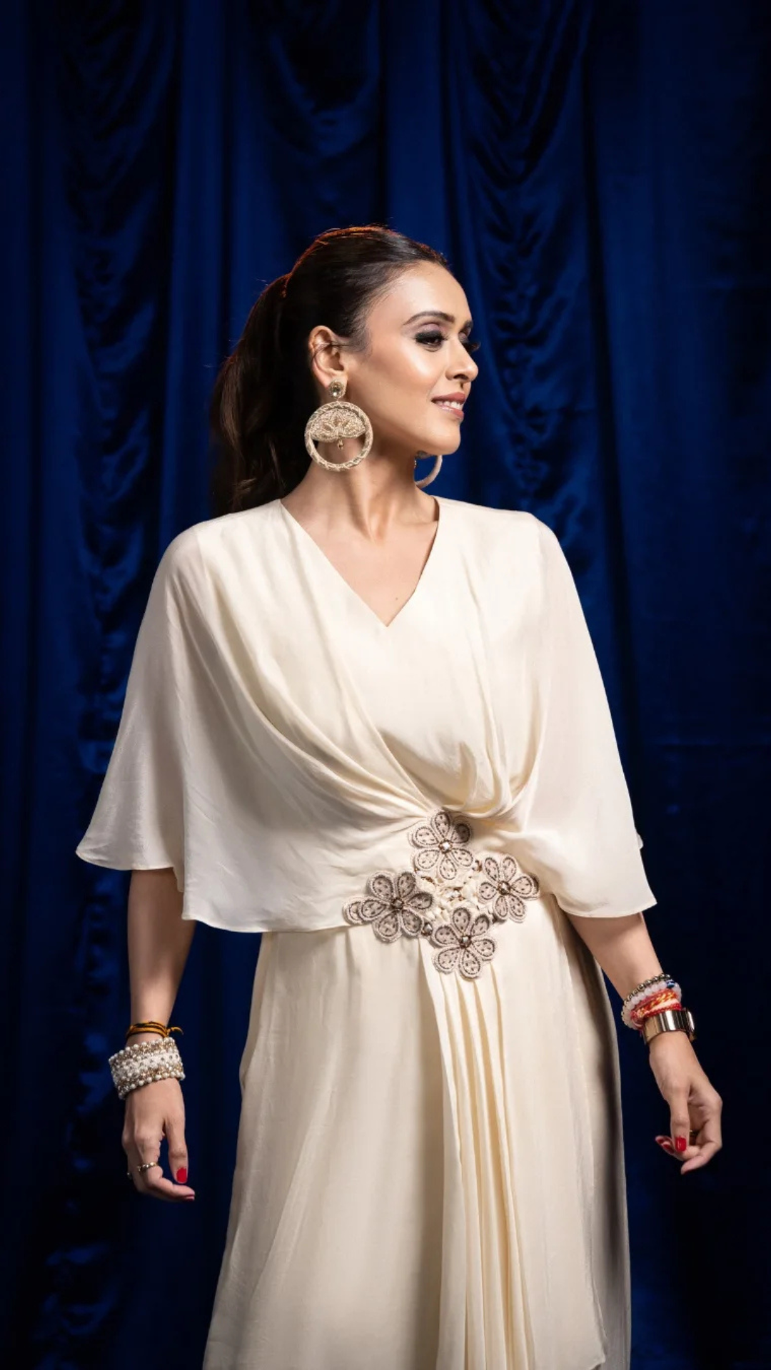 A Hrishitaa Bhatt wearing a cream draped wrap-style dress with a jeweled waist embellishment, standing against a dark blue draped backdrop. She is accessorized with large circular earrings and bracelets.