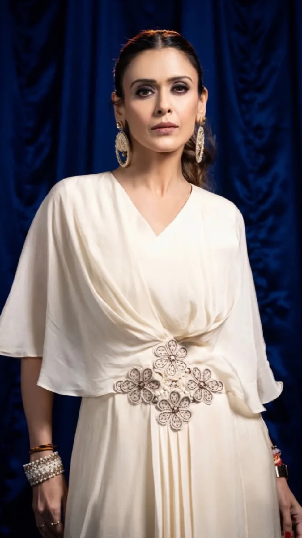 A Hrishitaa Bhatt wearing a cream-colored draped dress with a jeweled waist embellishment, standing against a dark blue velvet curtain backdrop.