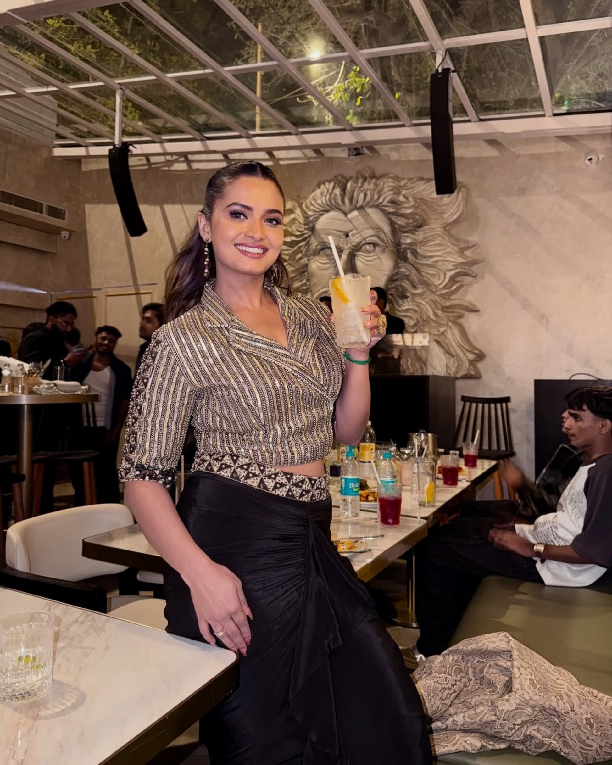 A Neelam Giri posing in a restaurant setting, wearing a metallic silver and black patterned wrap-style crop top with a black skirt, holding a drink.