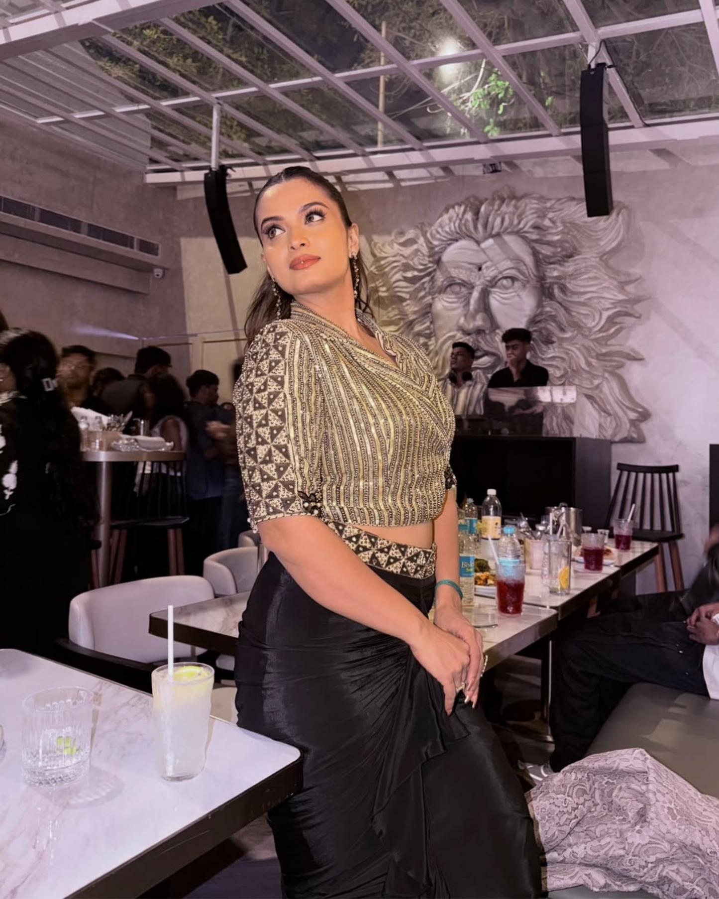 A Neelam Giri posing in a restaurant or event space, wearing a gold and black patterned short-sleeve crop top with a black satin skirt.
