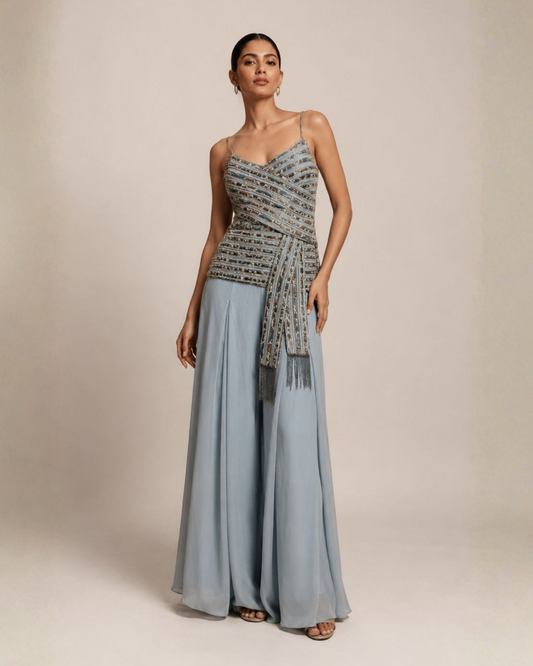 A woman models a sleeveless light blue formal gown with a heavily embellished beaded bodice in silver and blue tones, a matching beaded waist sash, and wide-leg flowing skirt, set against a plain beige studio background.