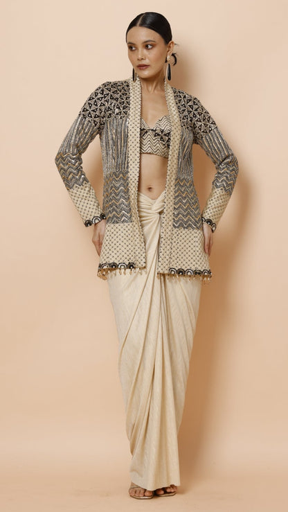 A woman modeling a traditional Indian outfit consisting of a cream-colored drape (sari) and a matching embroidered cardigan, set against a solid beige studio background.