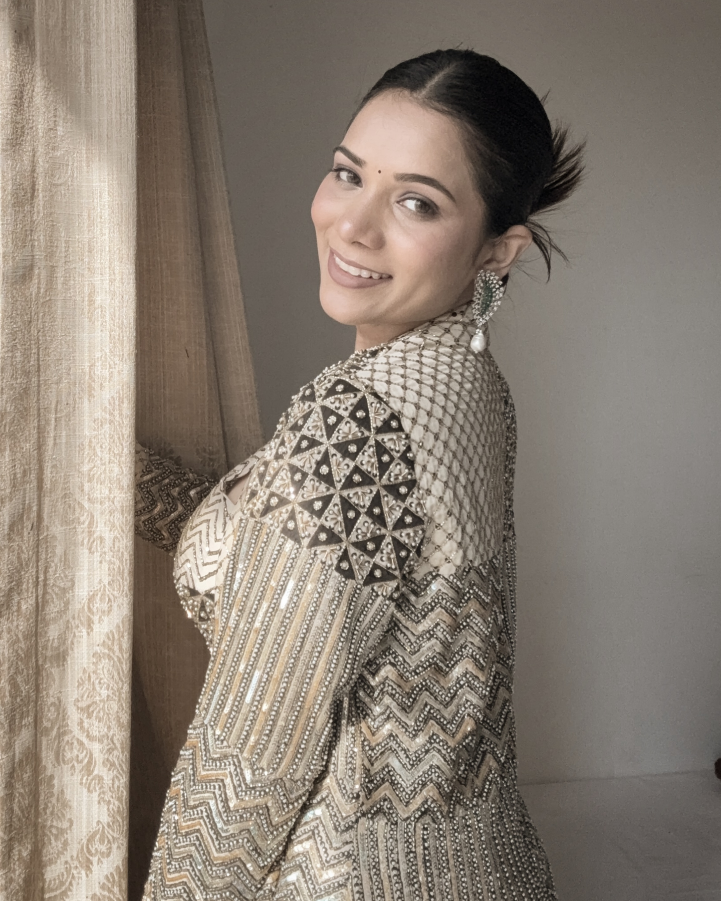 A Richa Archana wearing a heavily embellished beige and black traditional outfit with intricate silver beadwork and matching statement earrings, standing indoors next to a gold curtain.