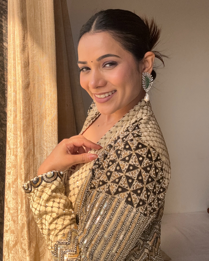 A Richa Archana wearing a heavily embellished beige and black traditional outfit with intricate silver beadwork and matching statement earrings, standing indoors next to a gold curtain.