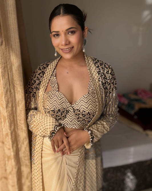 A Richa Archana wearing a beige and black patterned traditional outfit with a draped shawl, standing indoors near a curtain.