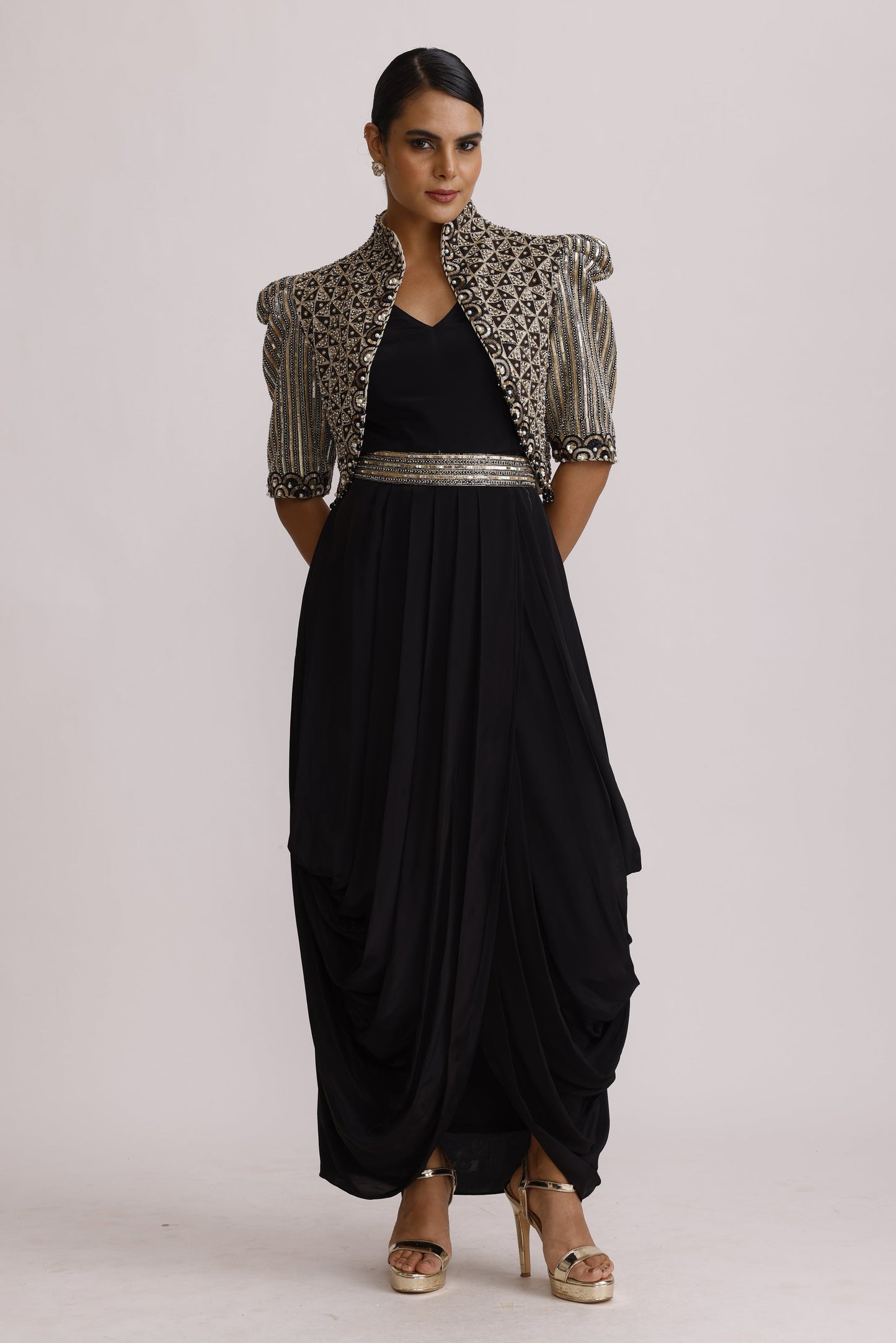 A woman modeling a black maxi dress with a silver beaded border, styled with a patterned silver and black jacket and gold high-heeled sandals, set against a plain white studio background.