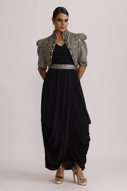 A woman modeling a black maxi dress with a silver beaded border, styled with a patterned silver and black jacket and gold high-heeled sandals, set against a plain white studio background.