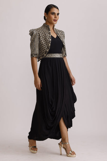 A woman modeling a black maxi dress with a draped, layered skirt, paired with a black and silver patterned shrug and beige high-heeled sandals, set against a plain light gray background.