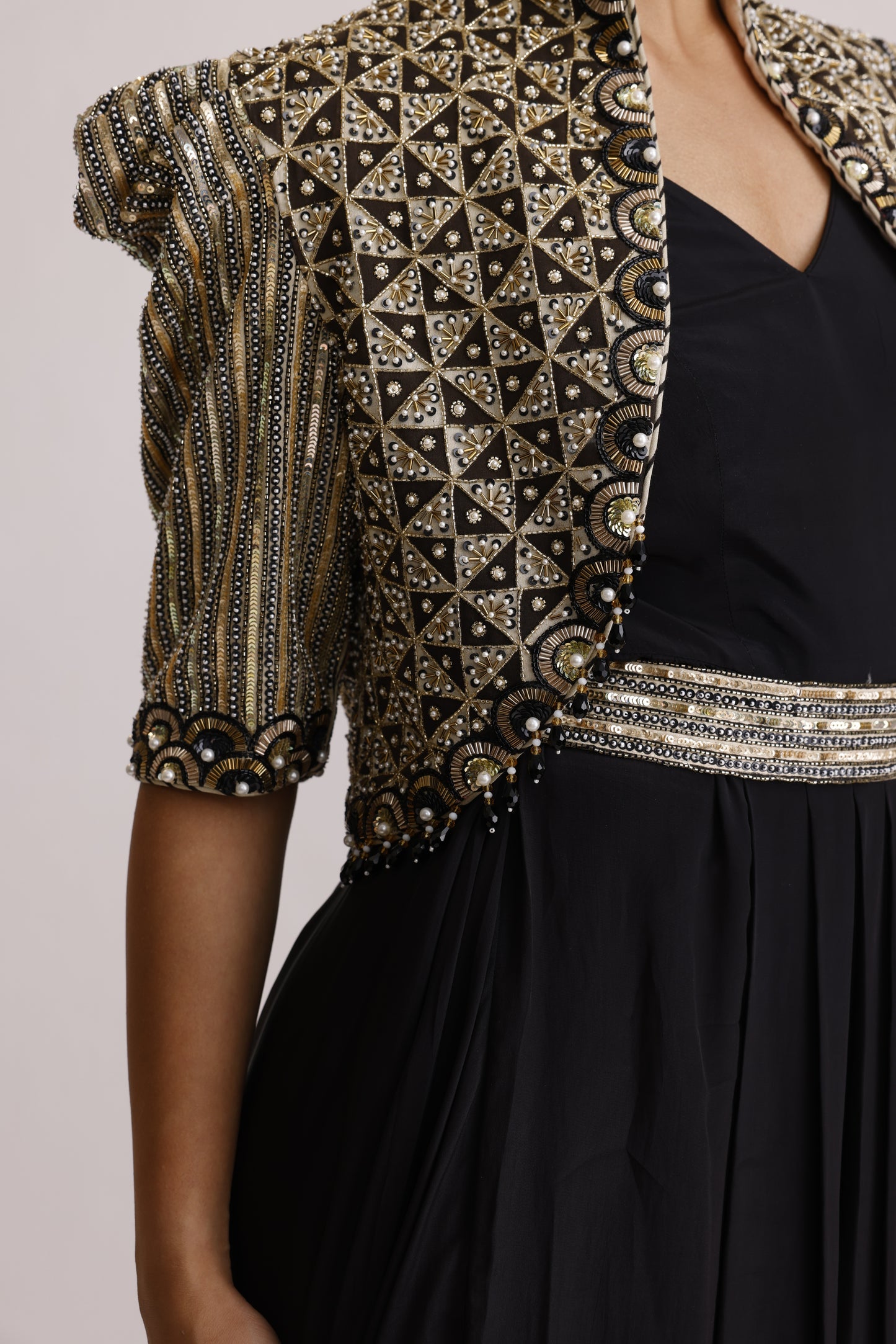 Close-up of a black garment with extensive metallic gold and silver geometric embroidery and beadwork on the sleeve and border, worn by a model.