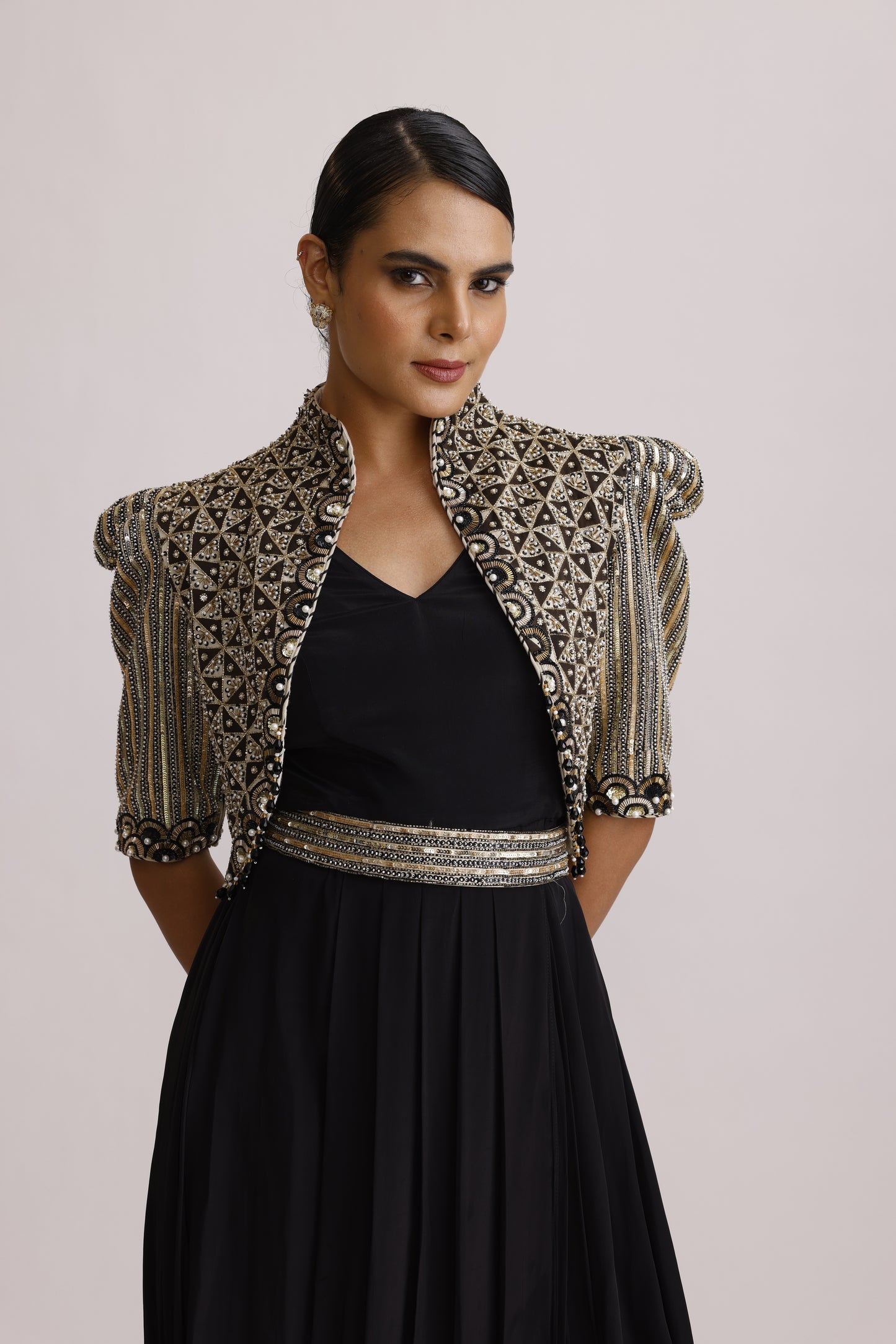 A woman wearing a black dress with a heavily embellished silver and gold embroidered jacket draped over her shoulders, photographed against a plain light background.
