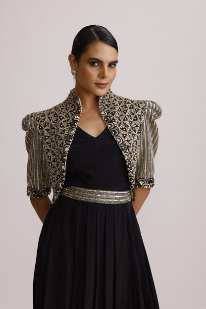 A woman wearing a black dress with a heavily embellished silver and gold embroidered jacket draped over her shoulders, photographed against a plain light background.