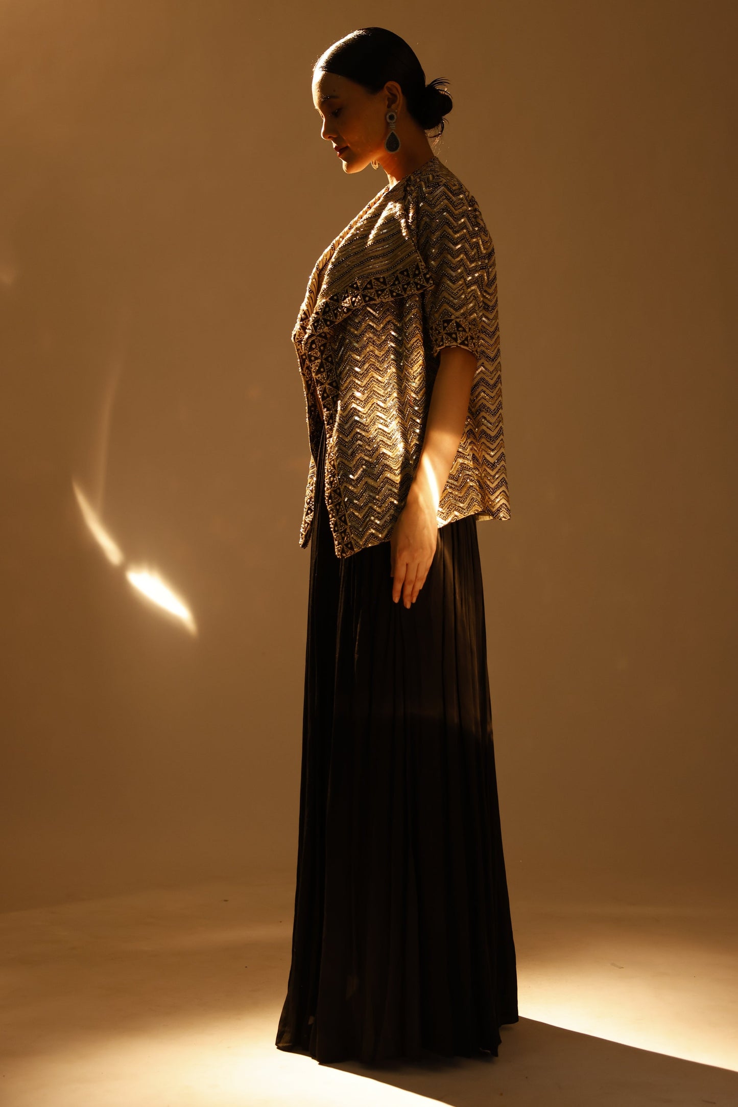 A model wearing a long black pleated skirt and a textured metallic silver top, posed against a warm, backlit beige wall.