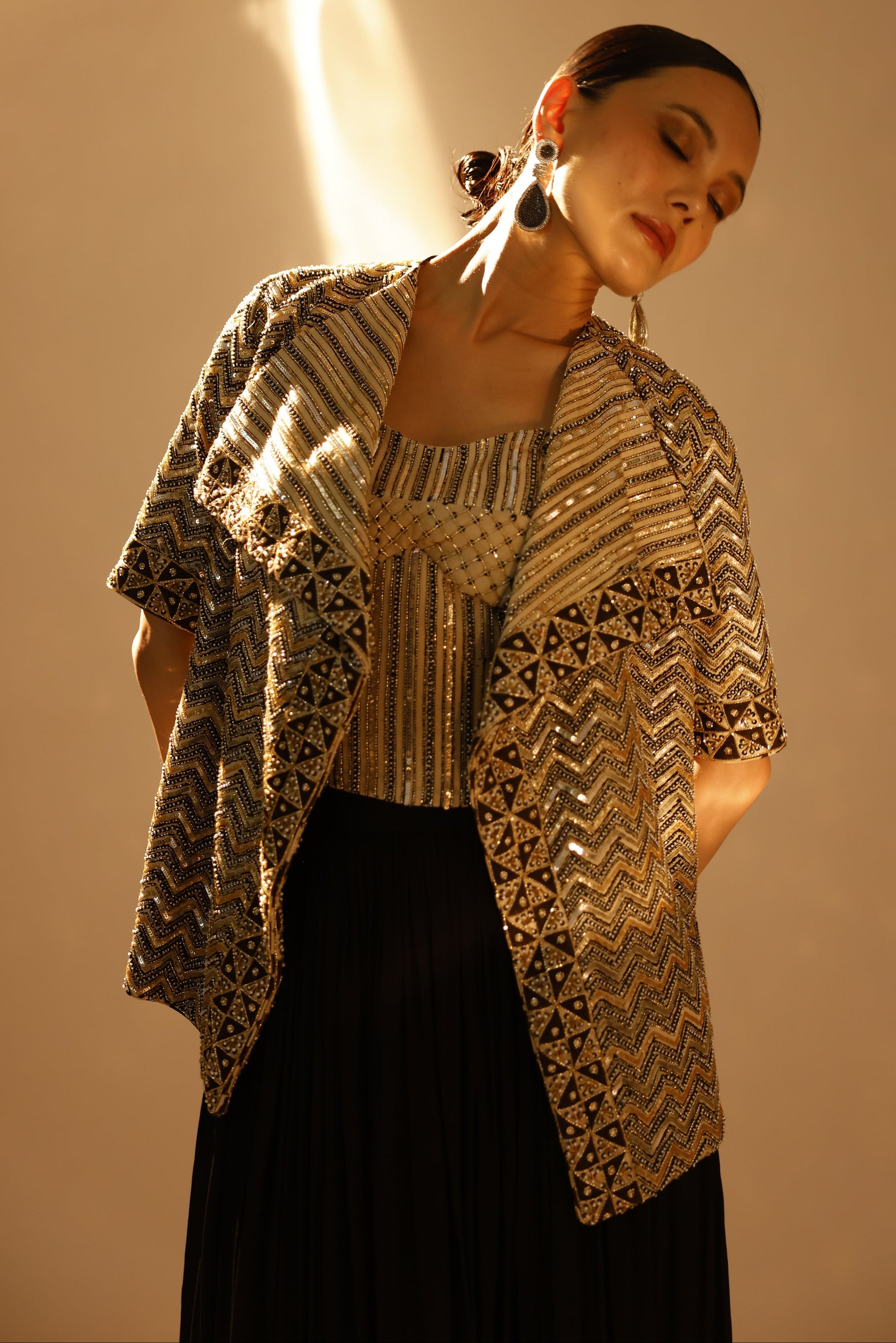 A woman wearing a black pleated skirt and a gold and black patterned embroidered jacket, posed against a warm beige background with dramatic side lighting.
