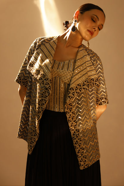 A woman wearing a black pleated skirt and a gold and black patterned embroidered jacket, posed against a warm beige background with dramatic side lighting.
