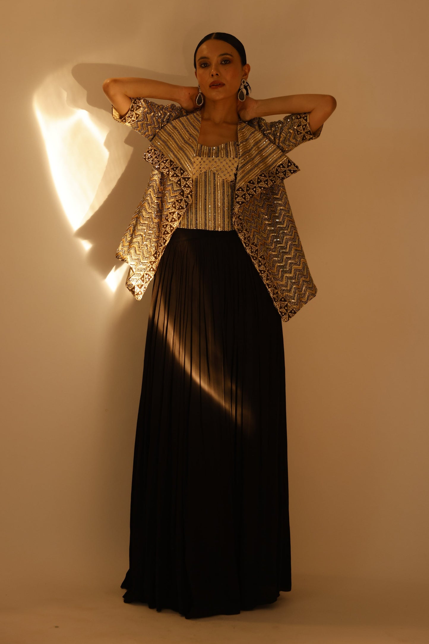 A woman modeling a long black pleated maxi skirt paired with a beige and black patterned ruffled jacket, standing against a warm beige wall with dramatic side lighting.