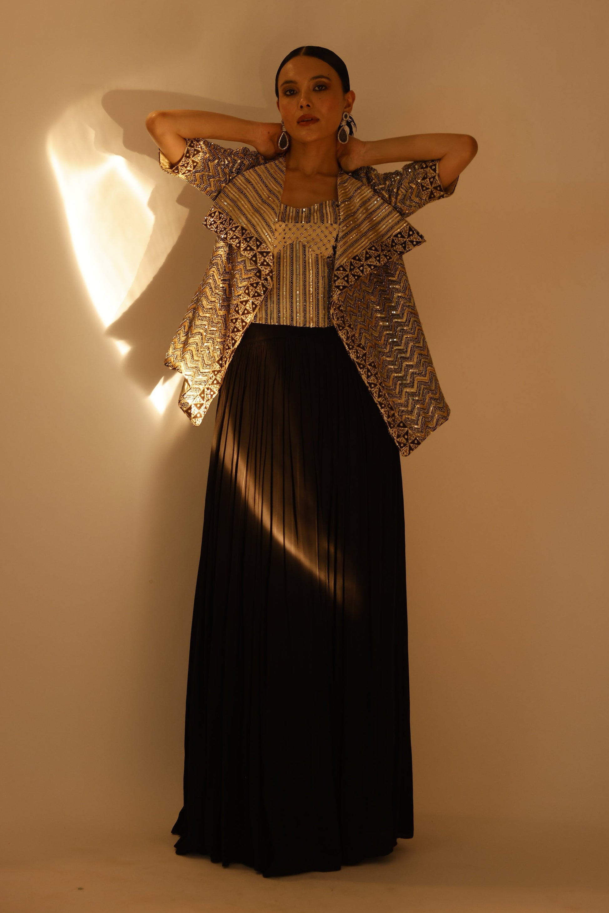 A woman modeling a long black pleated maxi skirt paired with a beige and black patterned ruffled jacket, standing against a warm beige wall with dramatic side lighting.