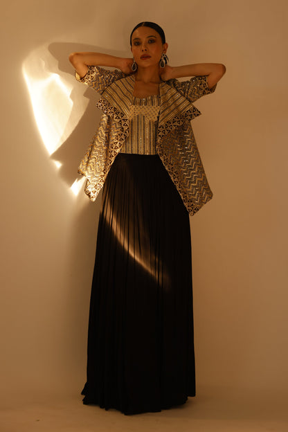A woman modeling a long black pleated maxi skirt paired with a beige and black patterned ruffled jacket, standing against a warm beige wall with dramatic side lighting.