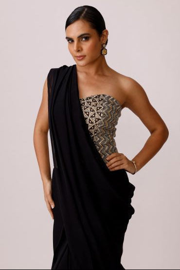 A woman modeling a black draped dress with a strapless bodice featuring intricate gold and silver geometric embroidery, posed against a solid light beige background.