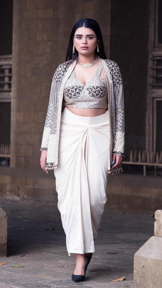 A Shreshta Iyer modeling a traditional Indian outfit featuring a cream-colored dhoti, a matching embroidered crop top, and a sheer embroidered cardigan, set against a rustic stone background.