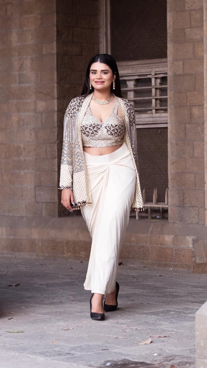 Shreshta Iyer wearing a traditional Indian outfit consisting of a cream-colored saree, a matching embroidered crop top, and a sheer embroidered cardigan, set against a stone wall background.