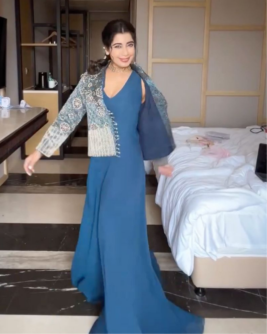 A Tanya Narang posing in a blue evening gown with a heavily embroidered teal and silver jacket draped over her shoulders, set in a modern hotel room.