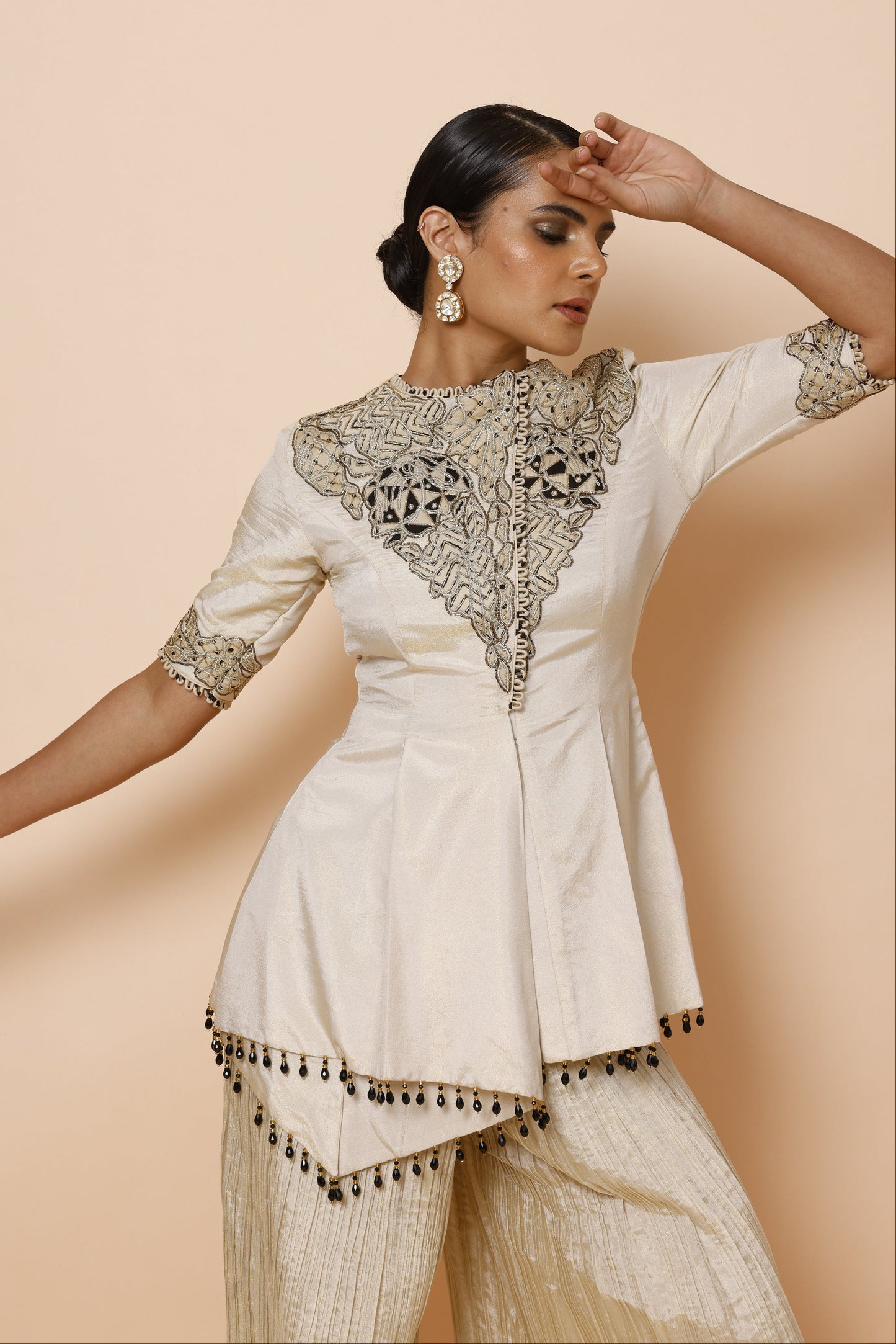 A woman wearing a cream-colored embroidered kurta with metallic gold and silver patterns on the chest and sleeve, paired with matching pleated trousers, posed dynamically against a solid beige background.