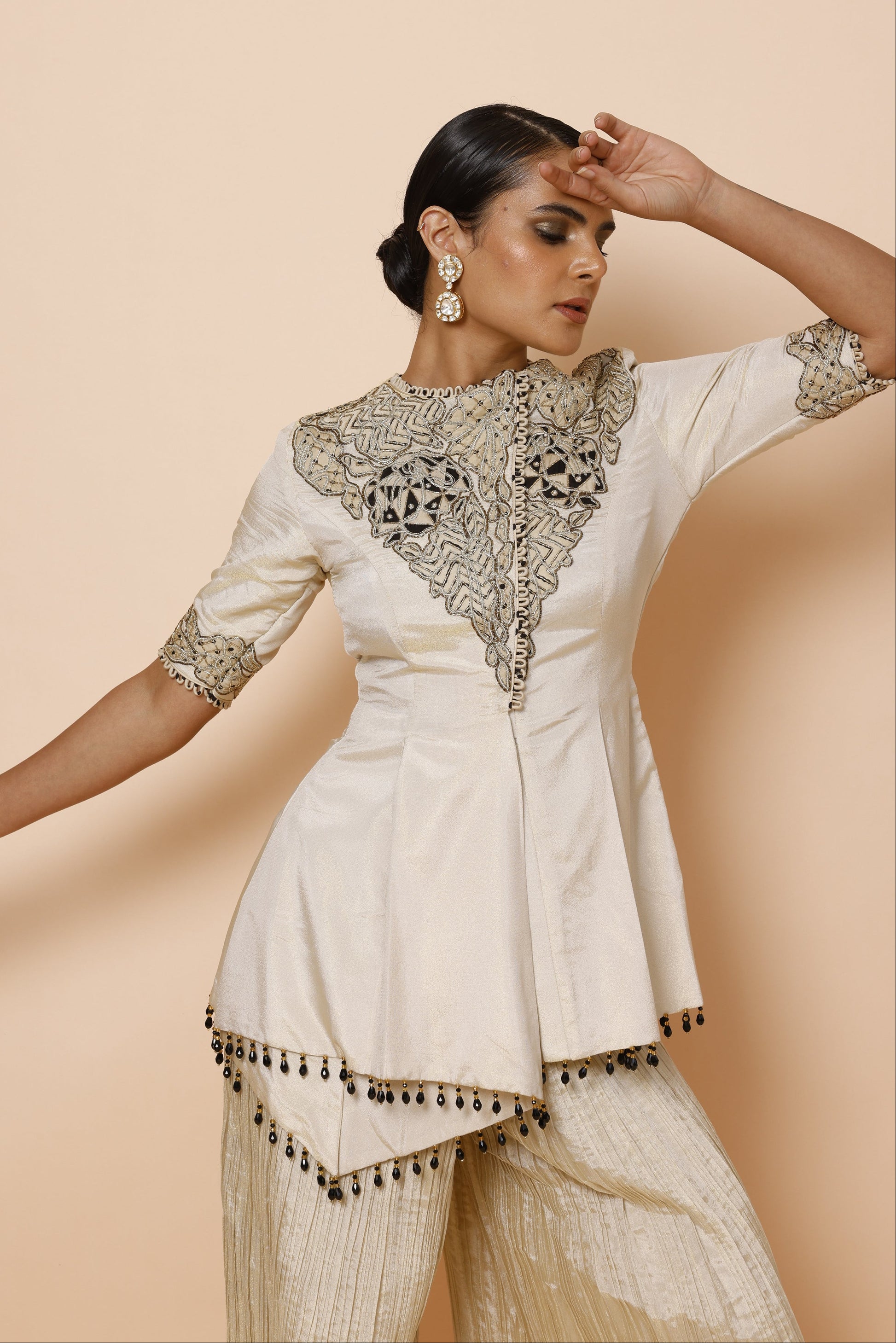 A woman wearing a cream-colored embroidered kurta with metallic gold and silver patterns on the chest and sleeve, paired with matching pleated trousers, posed dynamically against a solid beige background.