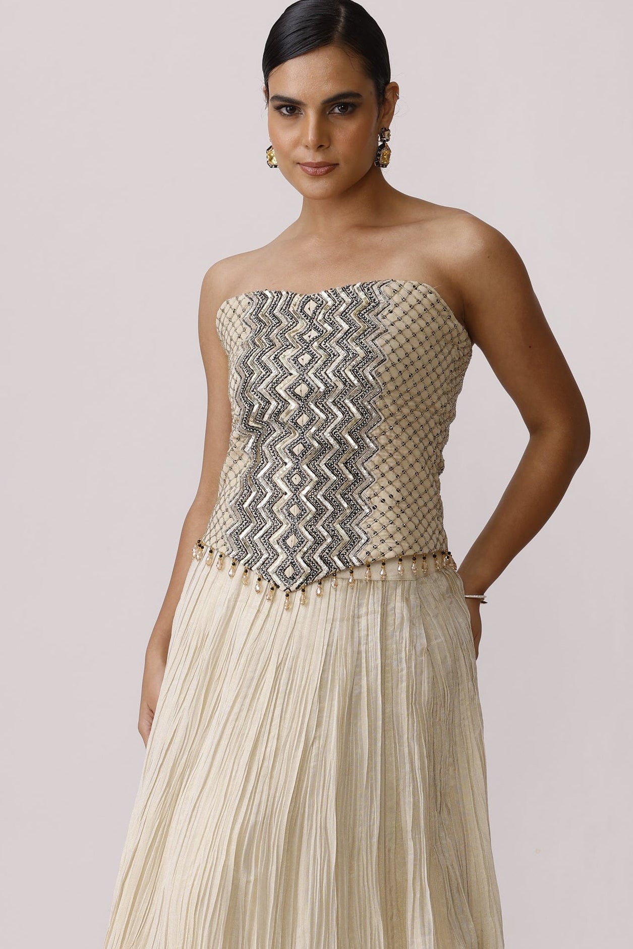 A model wearing a strapless floor-length gown with a pleated cream skirt and a textured, patterned bodice, photographed against a plain light background.