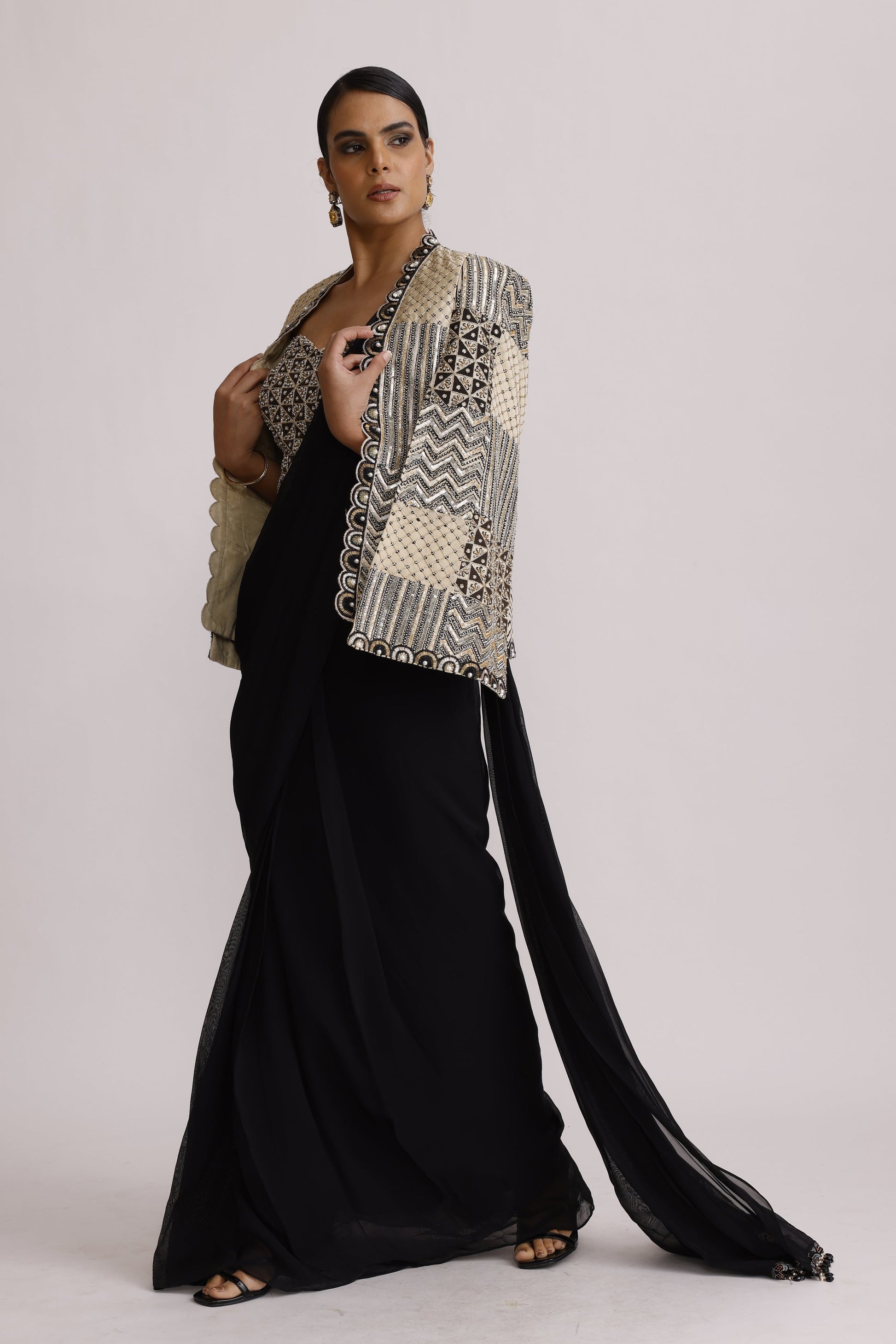 A model wearing a long black abaya with a wide, patterned beige and black shawl draped over her shoulder, standing against a plain light gray background.