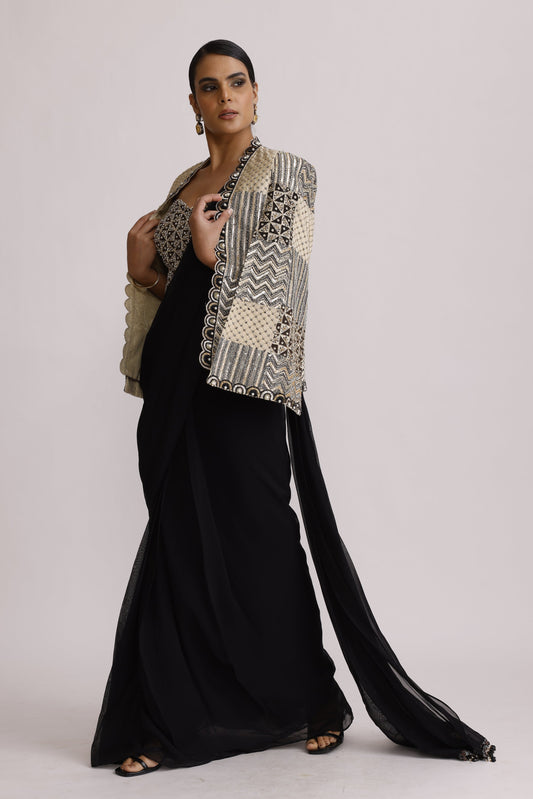 A model wearing a long black abaya with a wide, patterned beige and black shawl draped over her shoulder, standing against a plain light gray background.