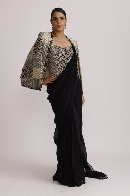 A model wearing a long black gown with a patterned beige and black draped overlay, posed against a plain light gray studio background.