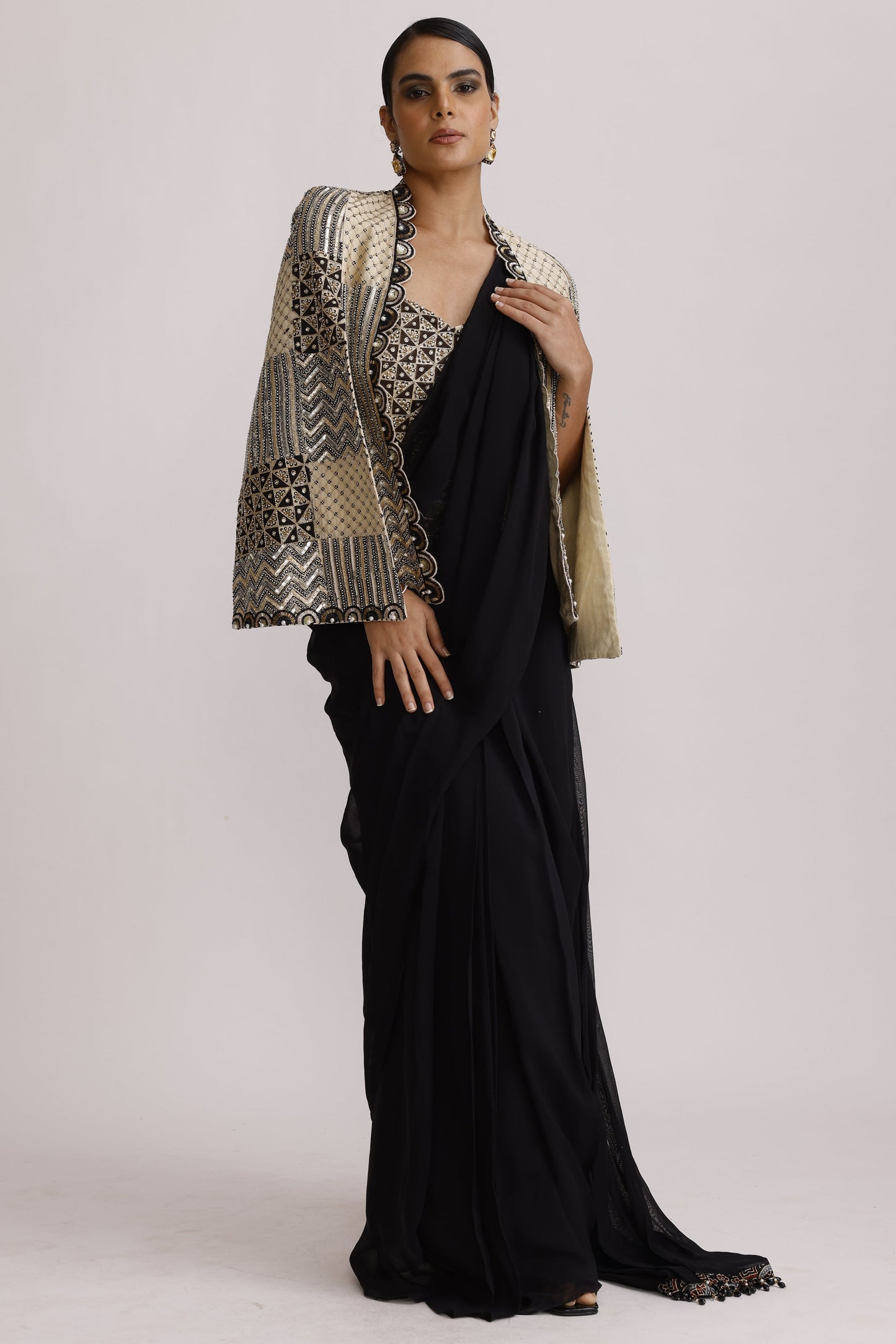 A model wearing a long black dress with a wide, patterned beige and black shawl draped over one shoulder, posed against a plain light gray studio background.