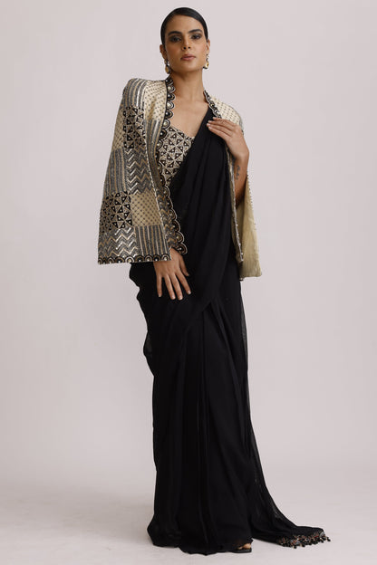 A model wearing a long black dress with a wide, patterned beige and black shawl draped over one shoulder, posed against a plain light gray studio background.