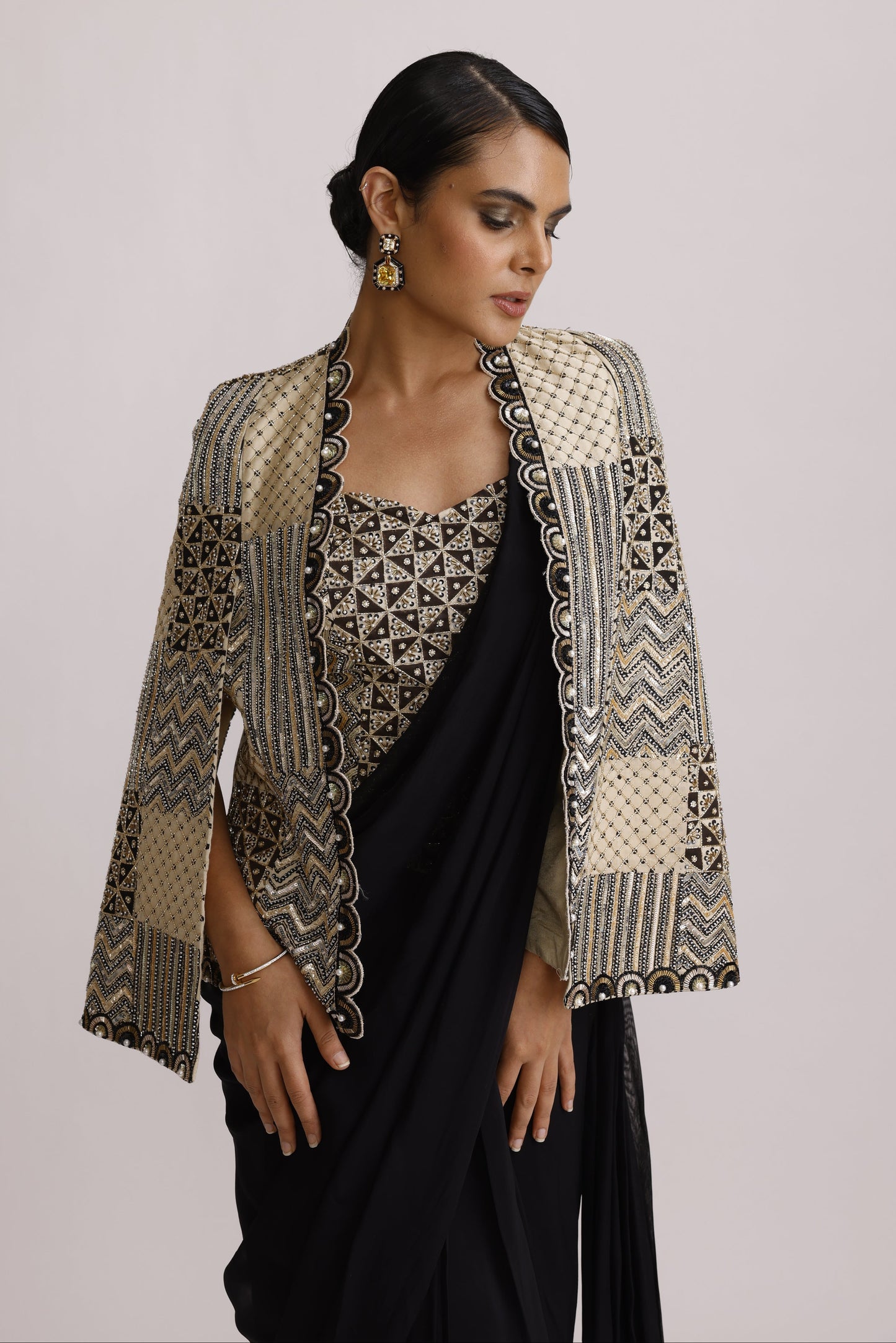 A woman wearing a black traditional outfit with a heavily embellished beige and black embroidered jacket featuring geometric patterns and scalloped edges, posed against a solid light gray background.